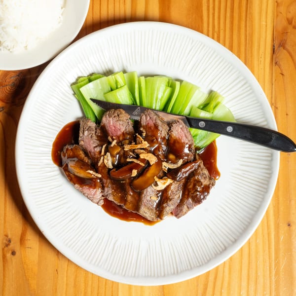 Sliced steak with mushroom gravy, crispy onions, and bok choy on a white plate with a side of rice.