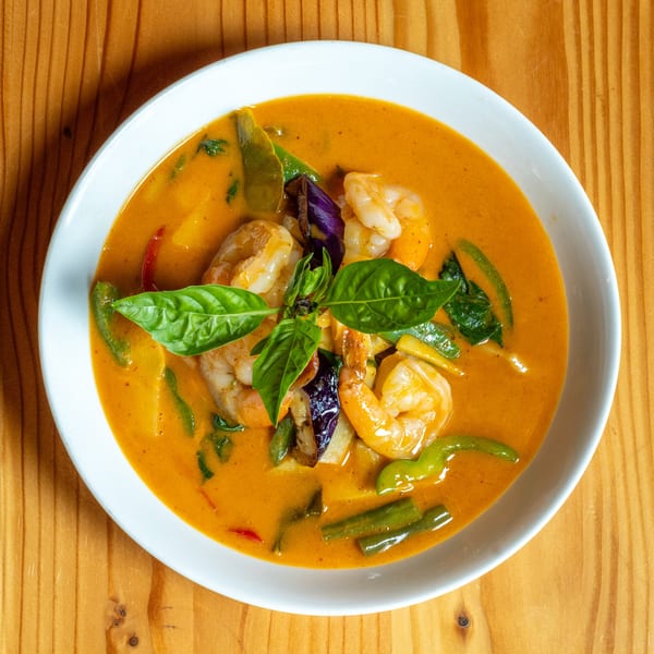 A white bowl of red curry with shrimp, green beans, eggplant, and basil on a wooden table.